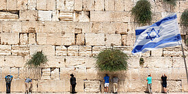 Die Klagemauer in Jerusalem mit betenden Menschen. Rechts im Vordergrund unscharf eine Israel-Flagge